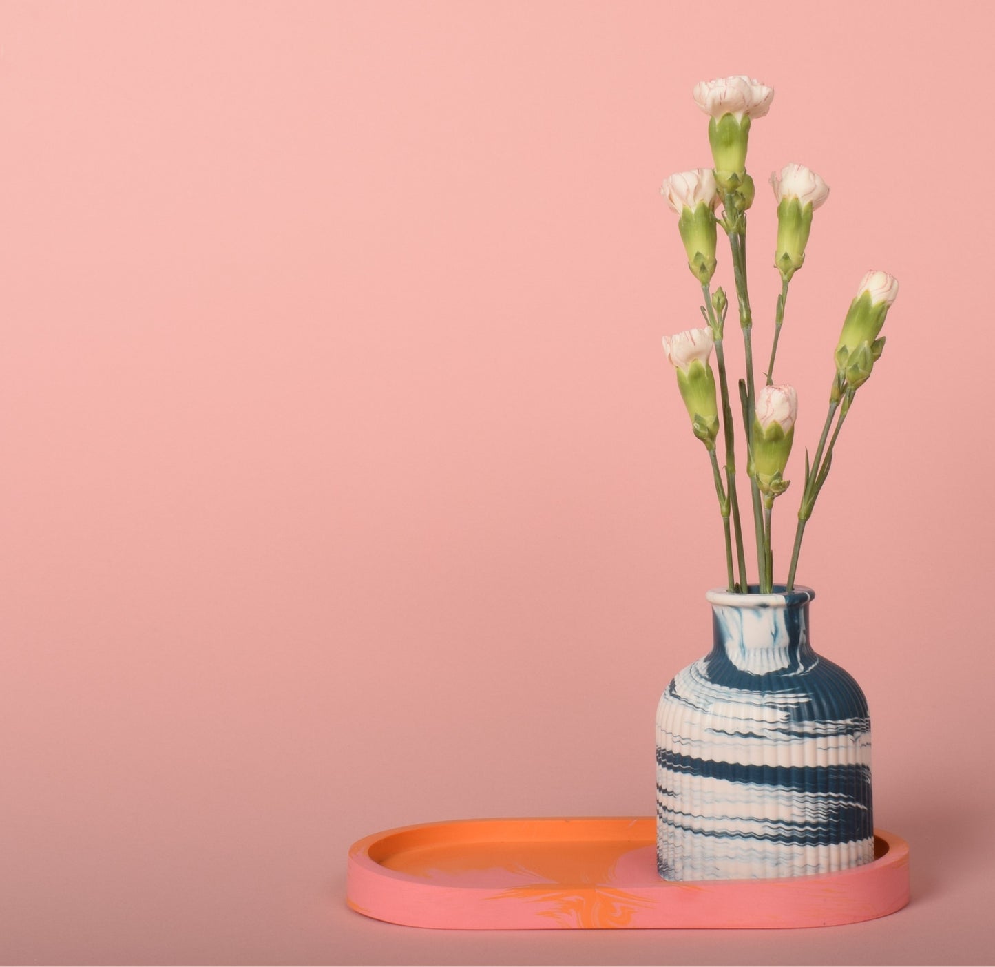 An oval marble effect pink and orange trinket tray holding a blue and white marble effect vase with flowers in it.