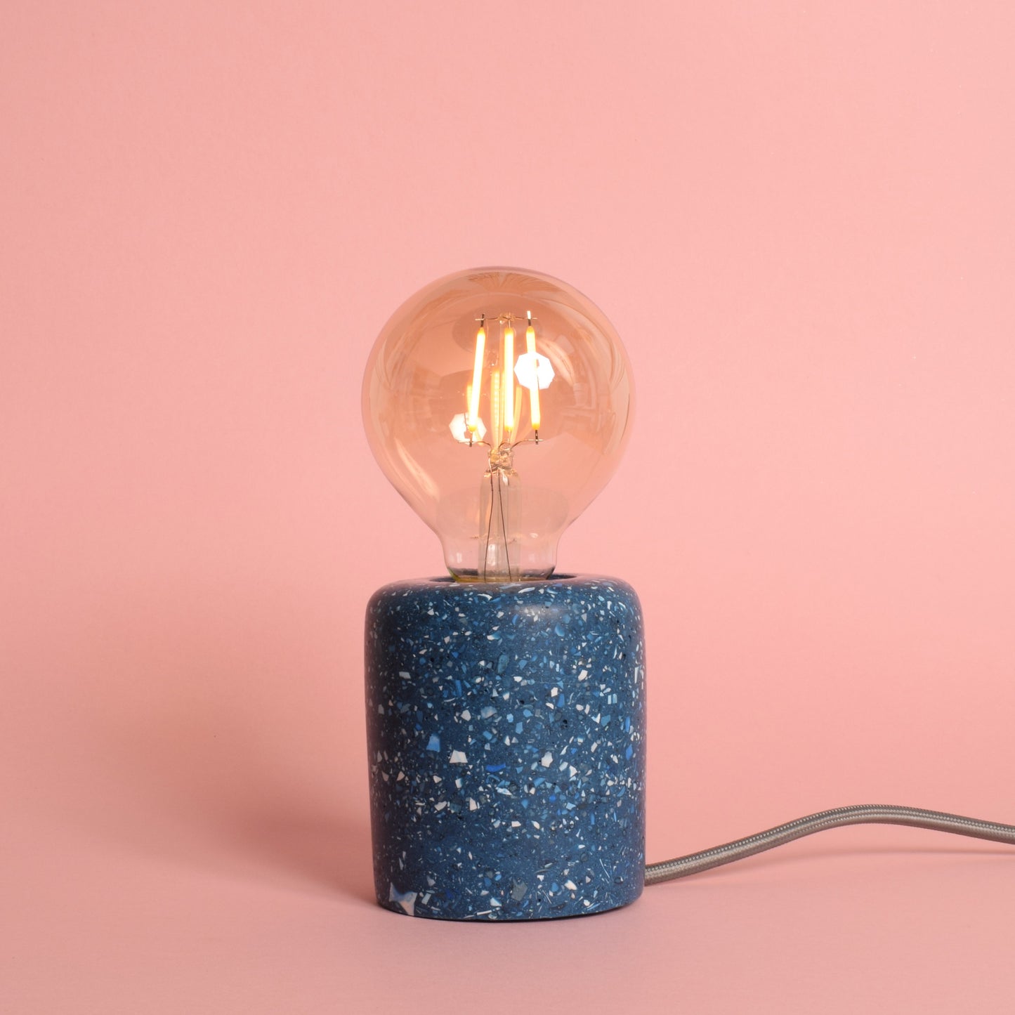 A blue rectangular e27 lamp base with blue and white terrazzo chips in it. The lamp has an exposed LED round bulb in it, and a grey wire coming out of the back. The lamp is turned on and gives out a warm amber glow.