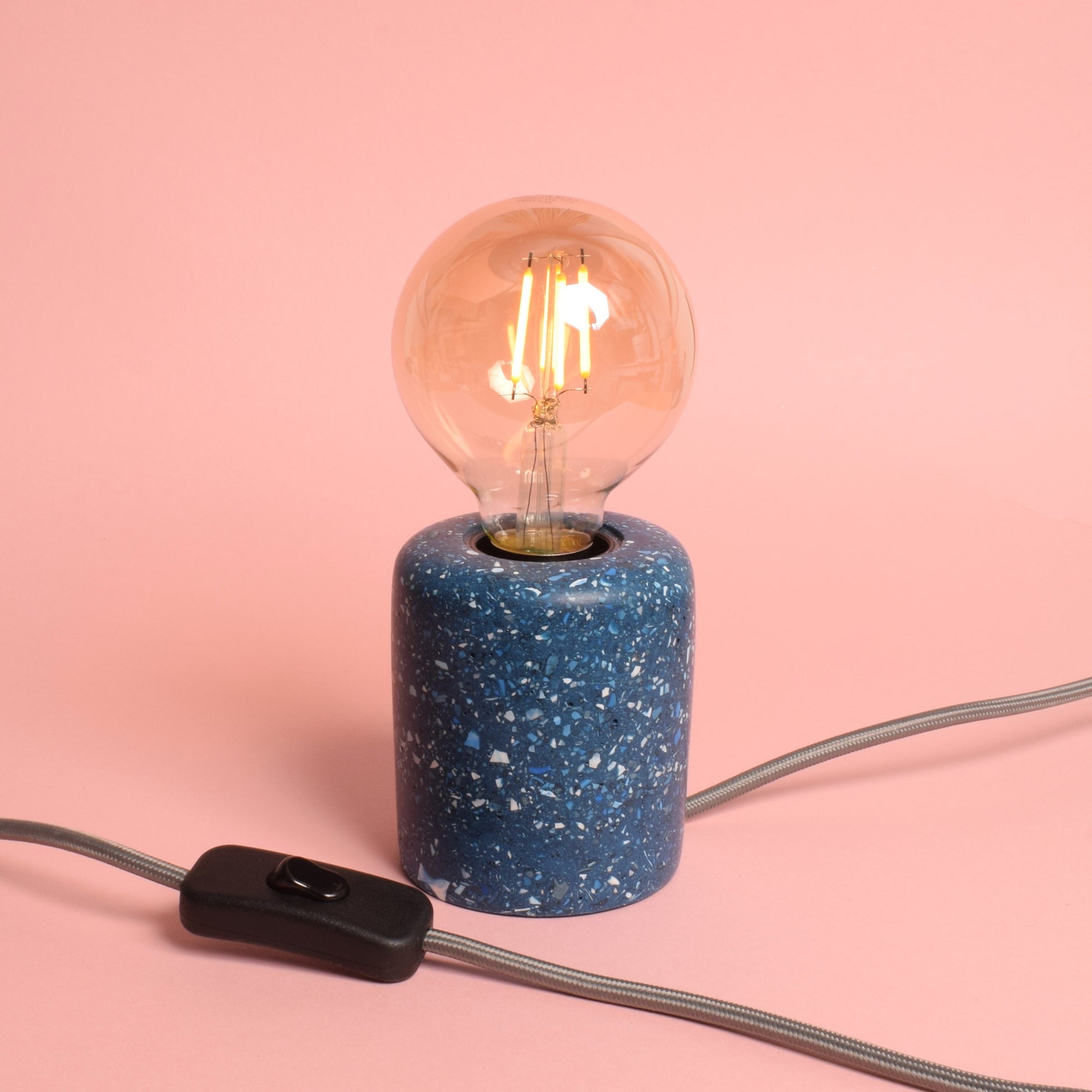 A blue rectangular e27 lamp base with blue and white terrazzo chips in it. The lamp has an exposed LED round bulb in it, and a grey wire coming out of the back, then back around the side, showing a black rocker switch. The lamp is turned on and gives out a warm amber glow. 