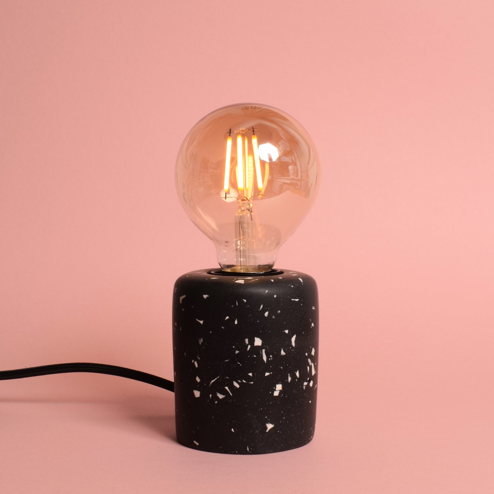 A black rectangular e27 lamp base with white terrazzo chips in it. The lamp has an exposed LED round bulb in it, and a black wire coming out of the back. The lamp is turned on and gives out a warm amber glow.