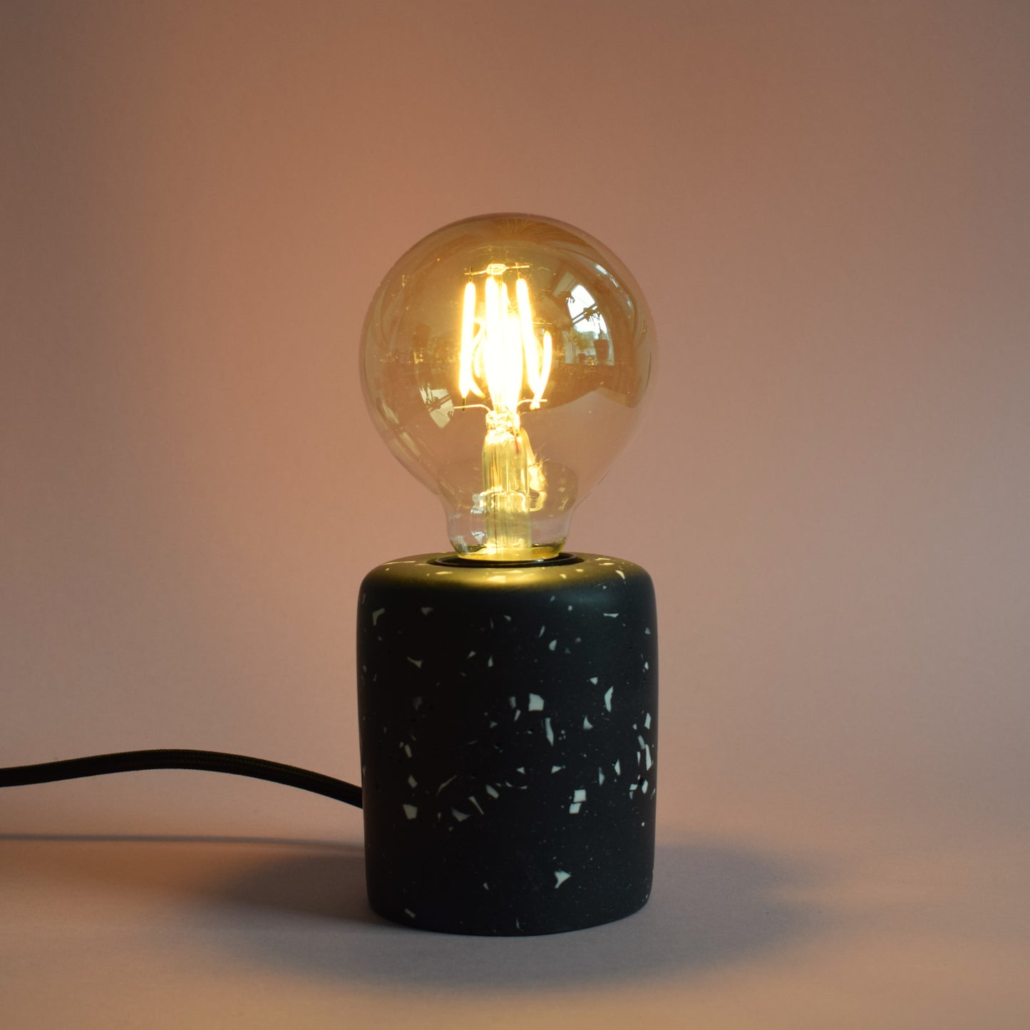 A black rectangular e27 lamp base with white terrazzo chips in it. The lamp has an exposed LED round bulb in it, and a black wire coming out of the back. The light is dim but the lamp is turned on and gives out a warm amber glow.