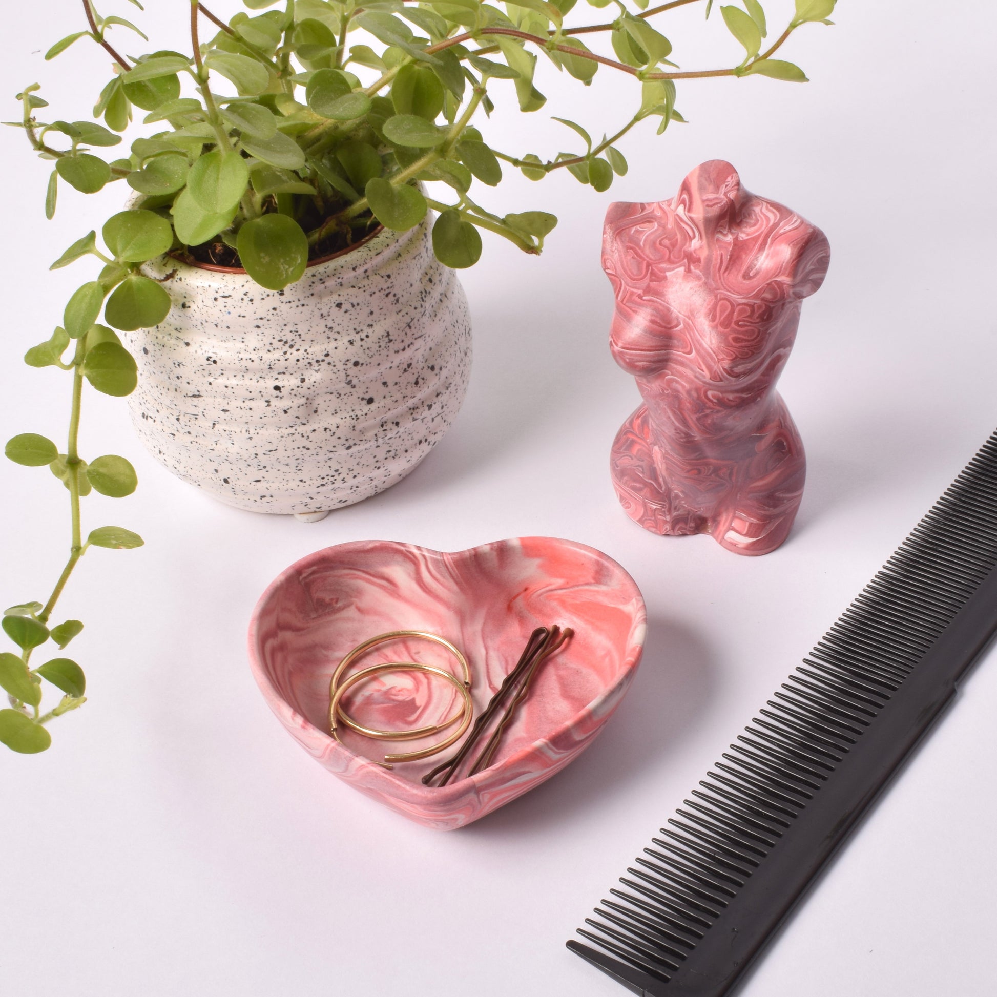 A heart shaped purple, pink and white trinket pot, holding earrings and bobby pins. There is a plant in the background, a purple female torso sculpture, and a comb.