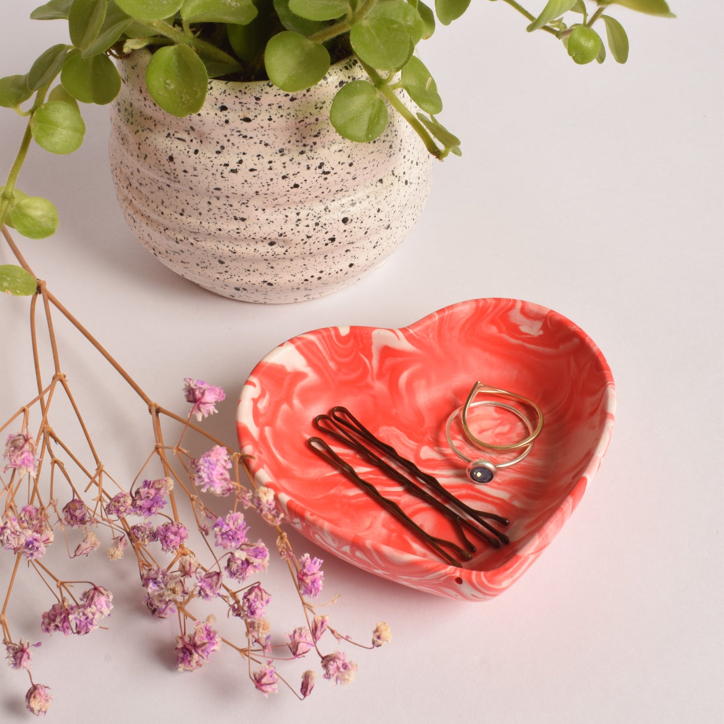 A heart shaped red and white trinket pot, holding rings and bobby pins. There is a plant in the background, and some dried flowers.