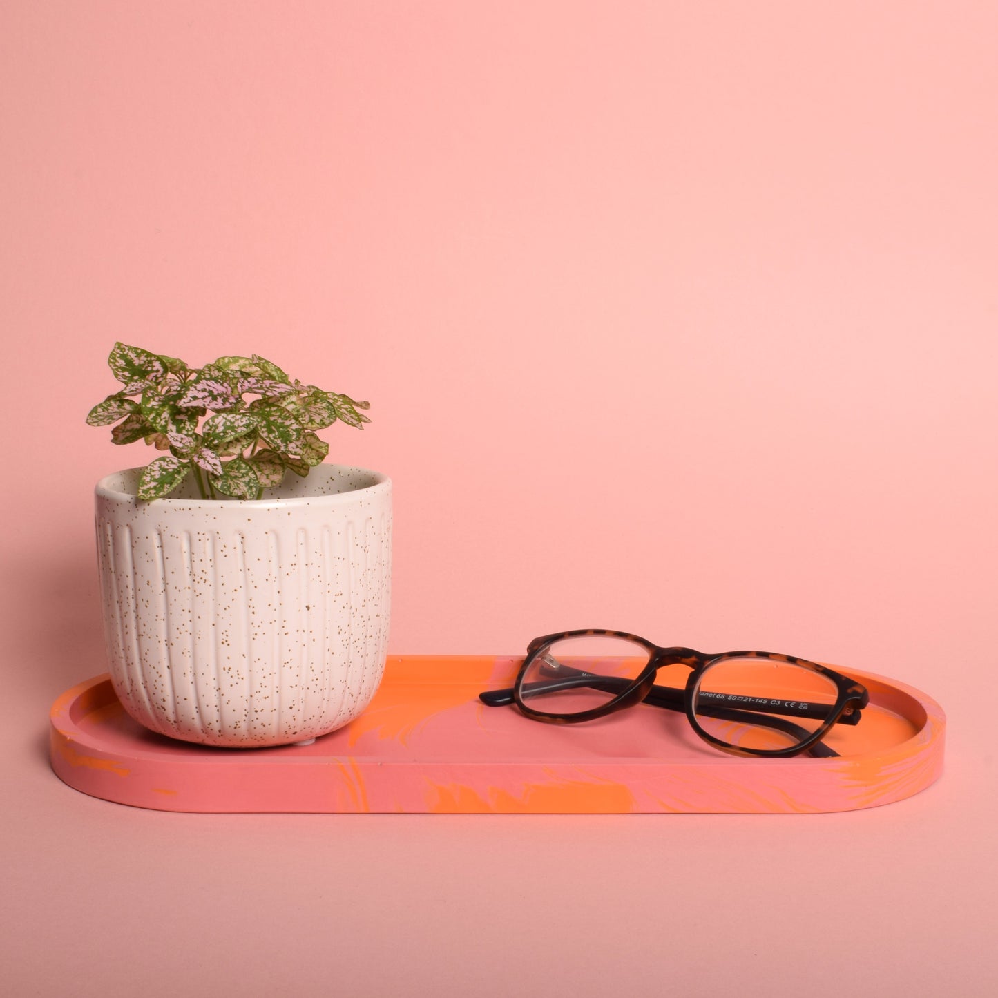 A large oval pink and orange marble effect trinket tray, holding a pair of glasses and a plant pot.