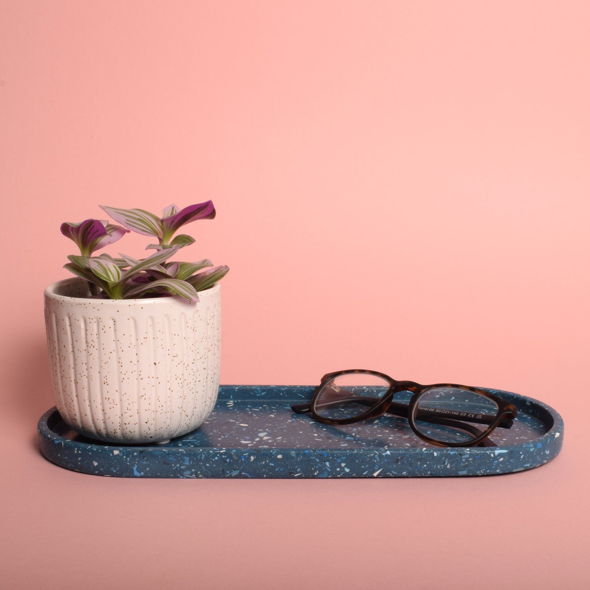 A large oval blue terrazzo trinket tray, holding a pair of glasses and a plant pot.