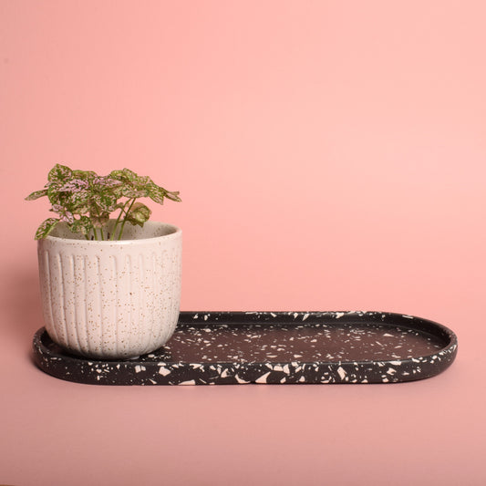 A large oval black trinket tray with white terrazzo chips in it. The tray is holding a plant pot with a plant in it.