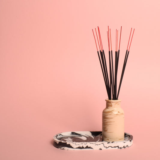 A black and white oval-shaped marble effect trinket tray with a beige marble effect vase on it. The vase is holding incense sticks