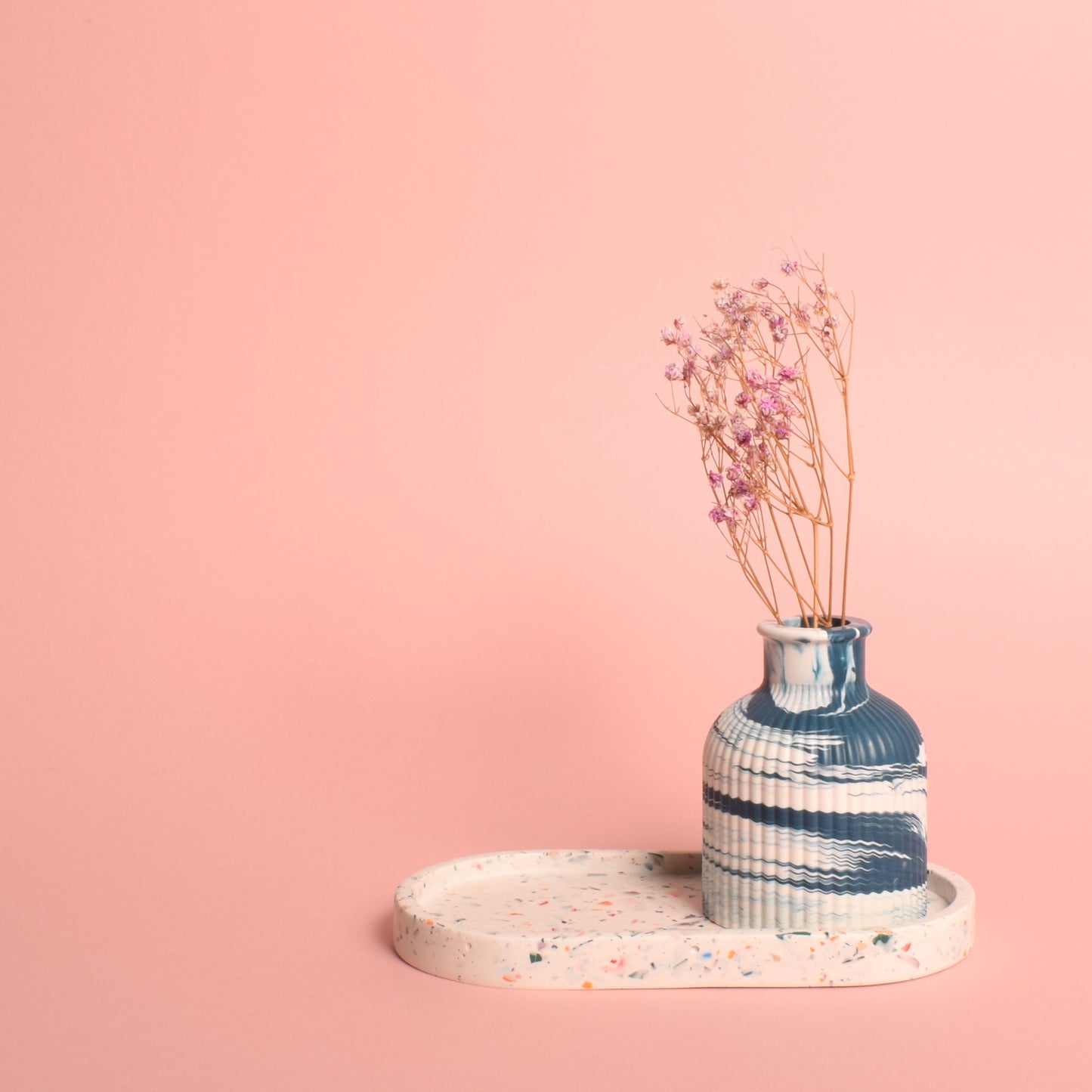 An oval-shaped white trinket tray with multi-coloured terrazzo chips in it. The tray is holding a blue and white marble effect ribbed vase with dried flowers in it.