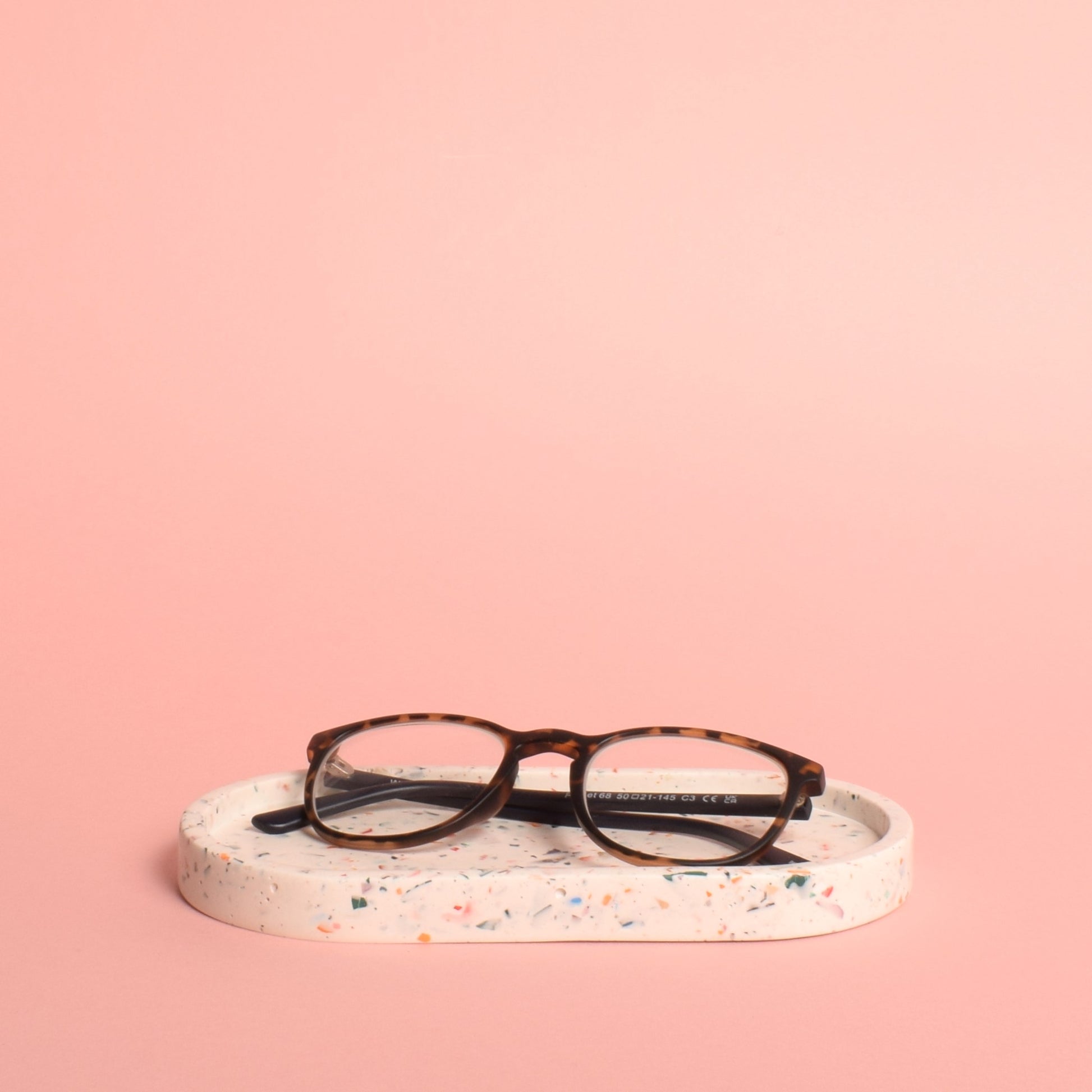 An oval-shaped white trinket tray with multi-coloured terrazzo chips in it. The tray is holding a pair of glasses.