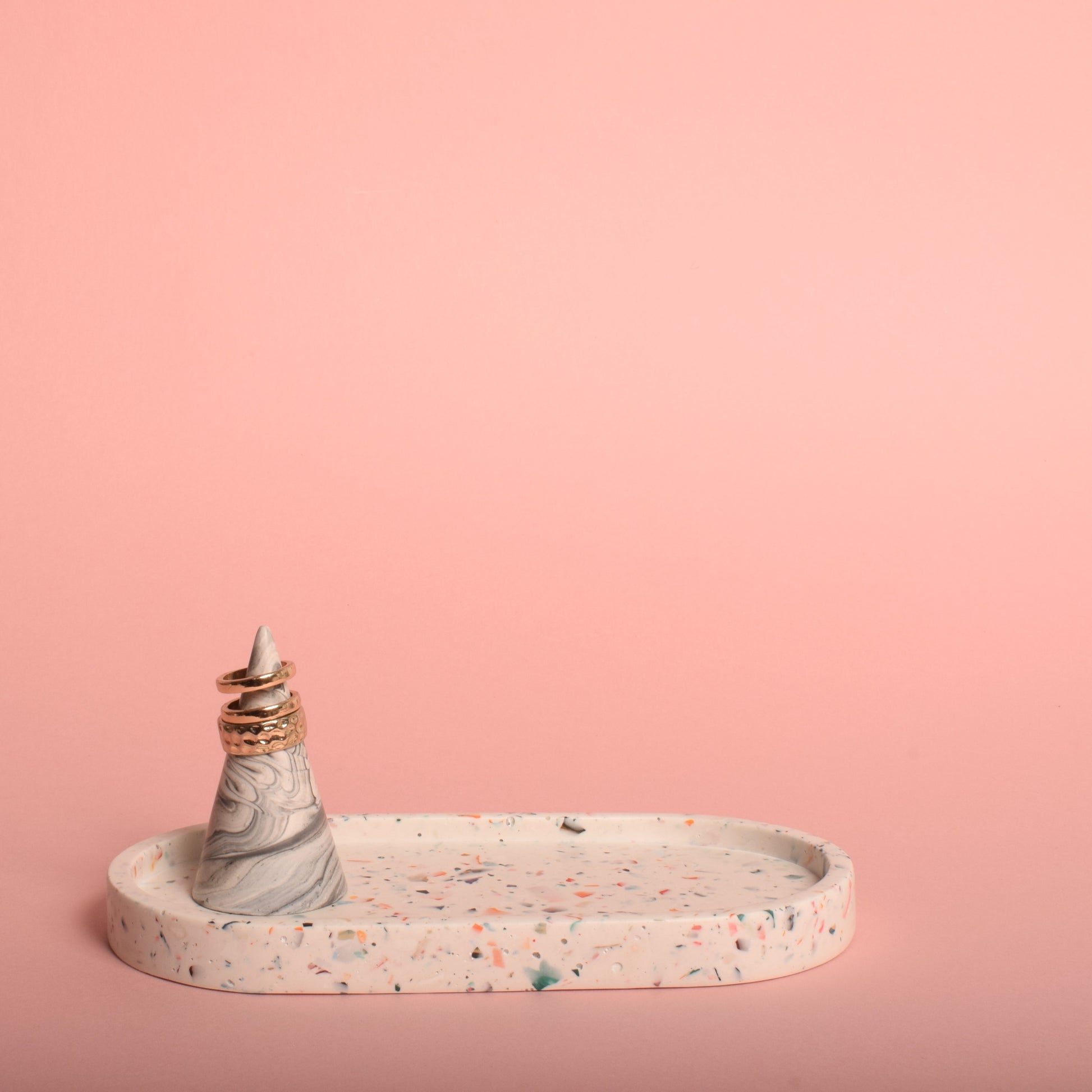 An oval-shaped white trinket tray with multi-coloured terrazzo chips in it. The tray is holding a grey and white ring holder with gold rings on it.