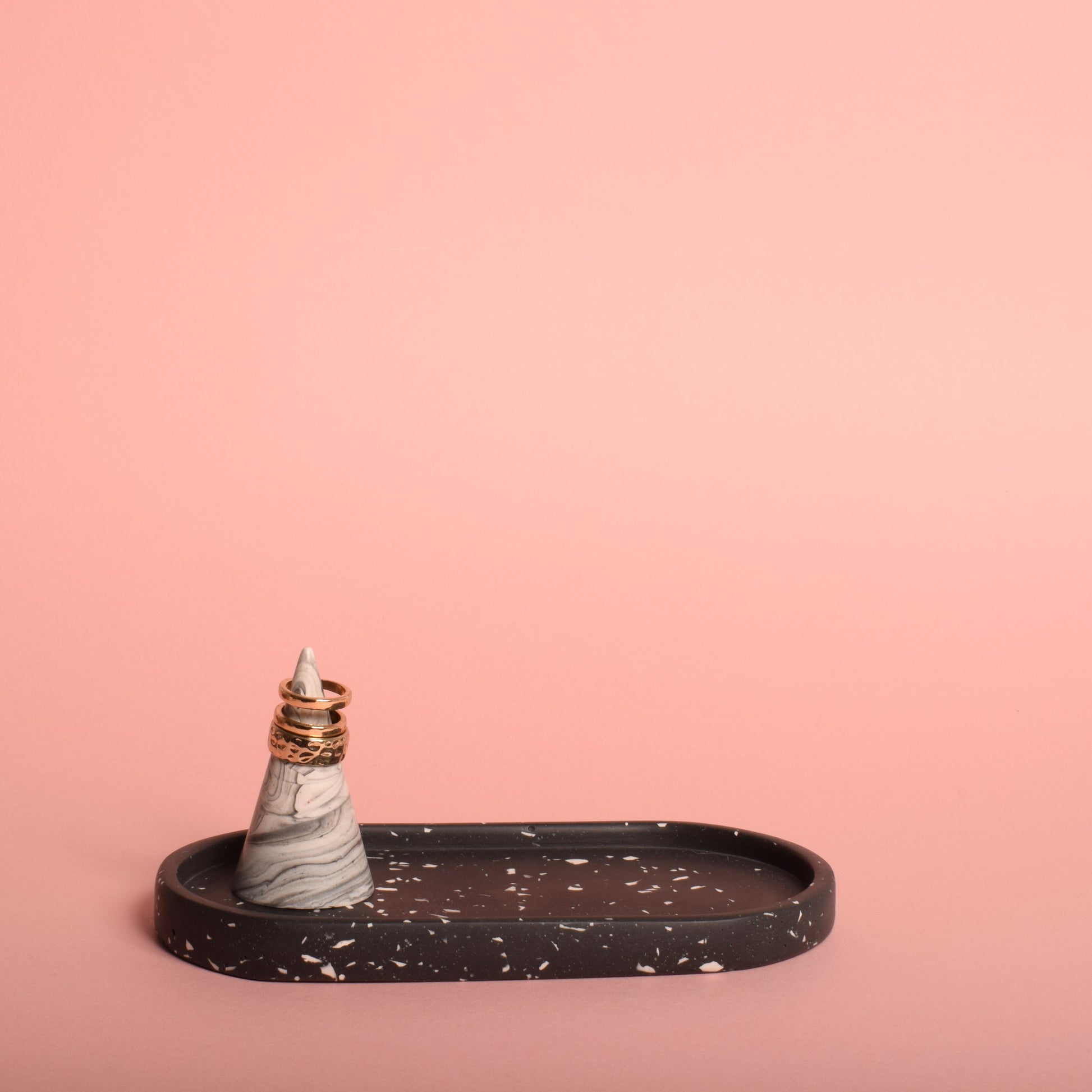 An oval-shaped black trinket tray with white terrazzo chips. The trinket tray is holding a grey and white ring holder, with gold rings on it.