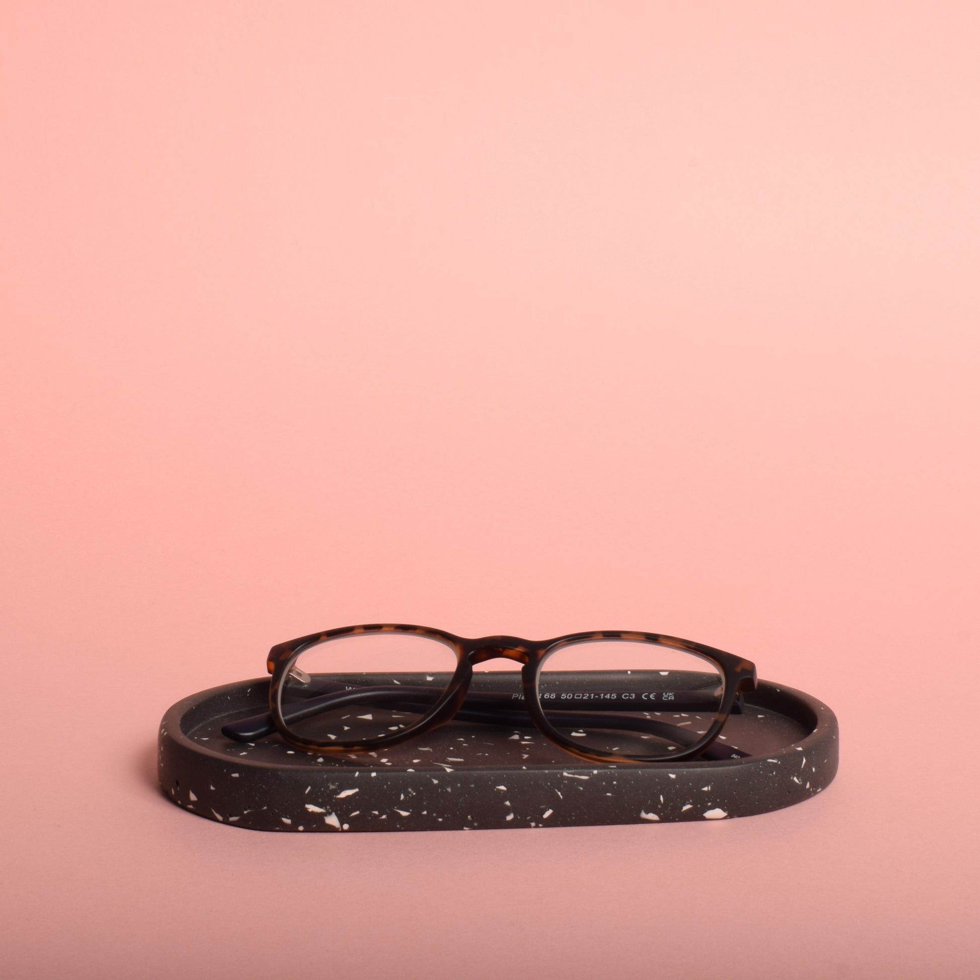 An oval-shaped black trinket tray with white terrazzo chips. The trinket tray is holding a pair of glasses.