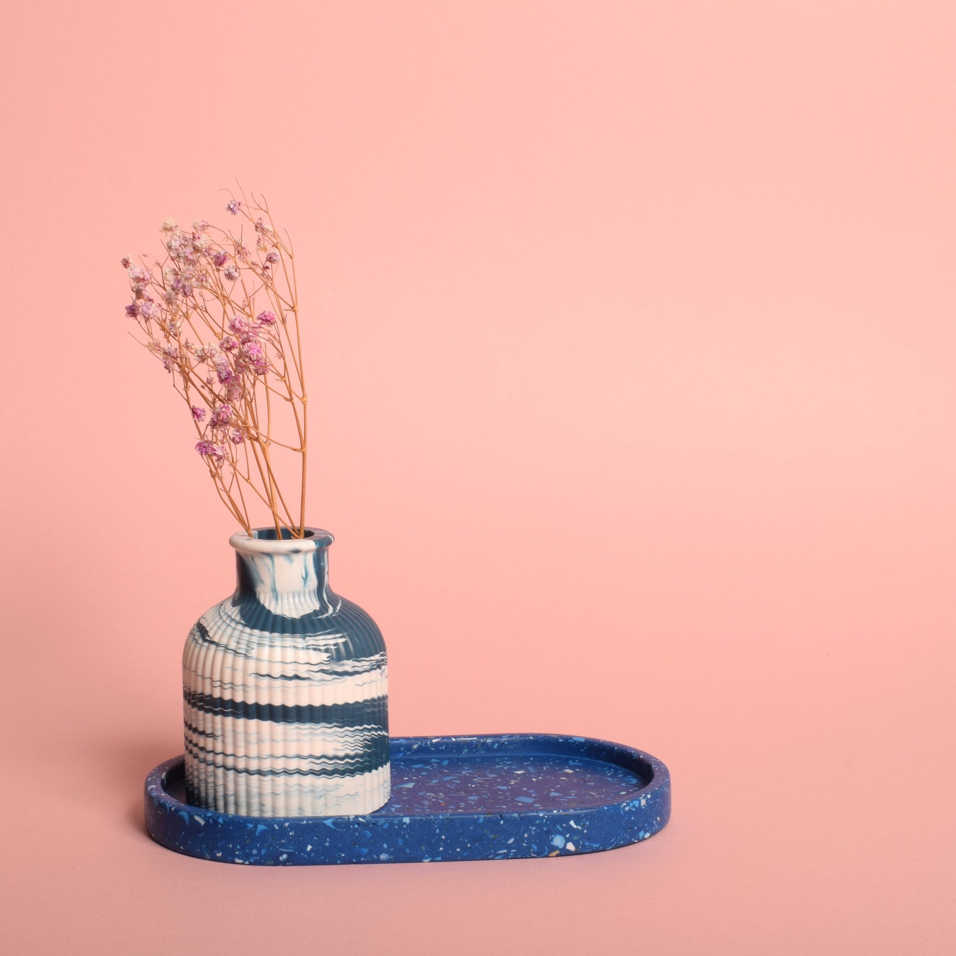 A blue oval trinket tray with blue and white terrazzo chips in it. The tray is holding a blue and white vase with dried flowers in it.