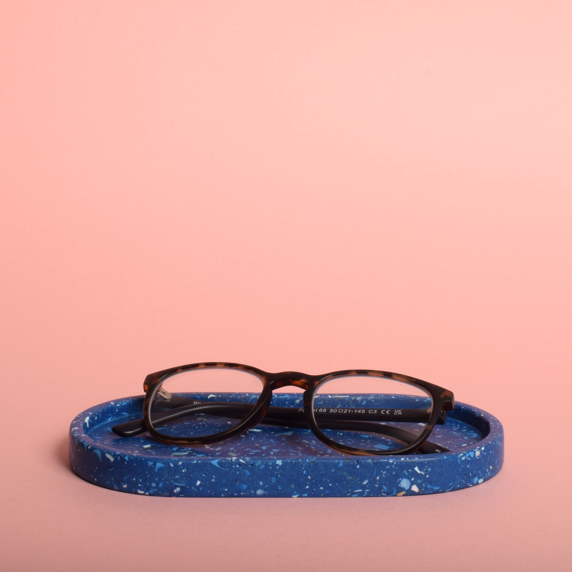 A blue oval trinket tray with blue and white terrazzo chips in it. The tray is holding a pair of glasses.