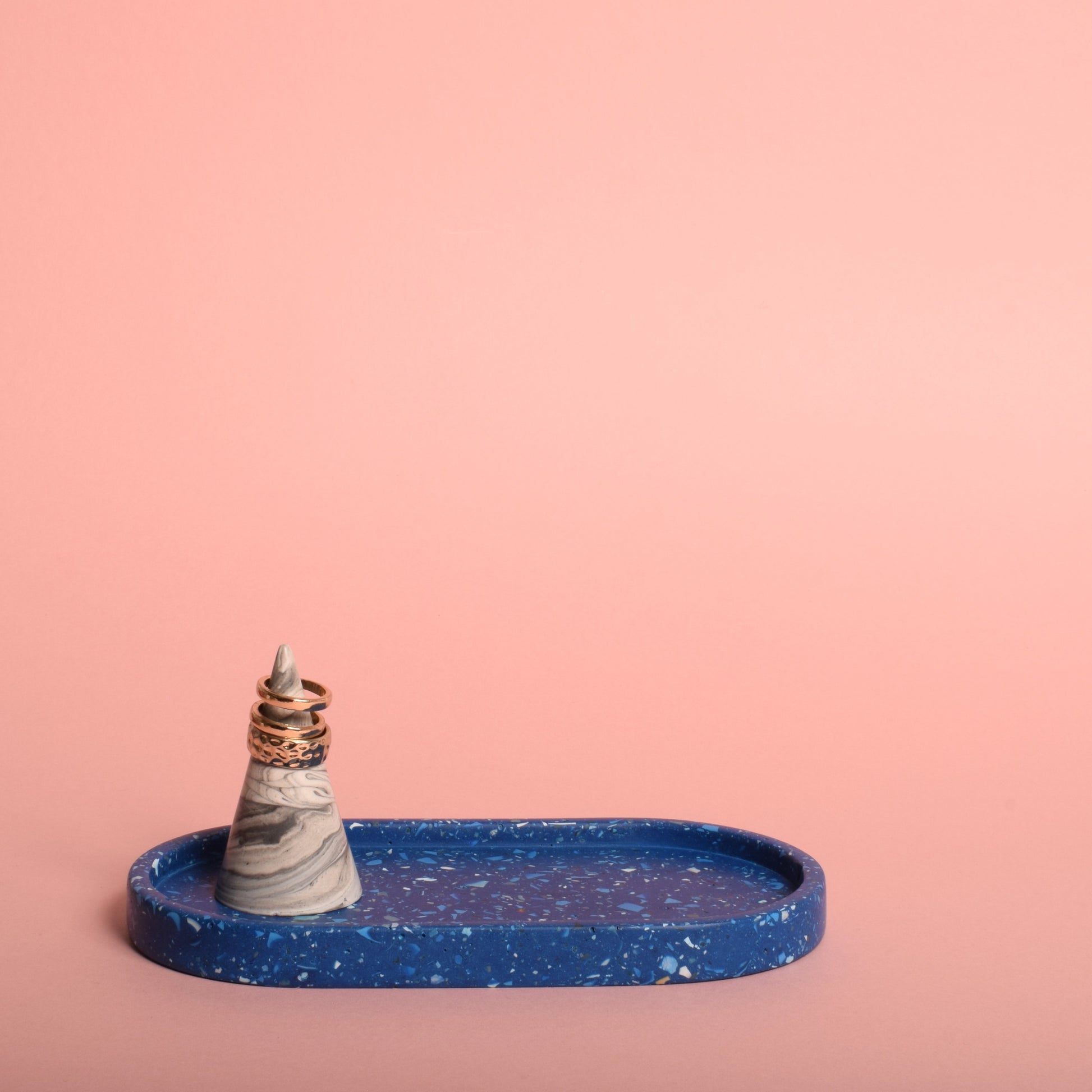 A blue oval trinket tray with blue and white terrazzo chips in it. The tray is holding a grey and white ring holder, which is holding gold rings.