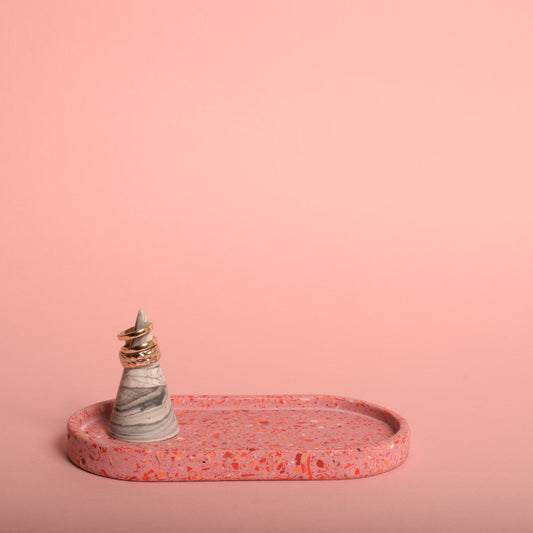 An oval pink terrazzo trinket tray holding a grey and white ring holder with gold rings on it.