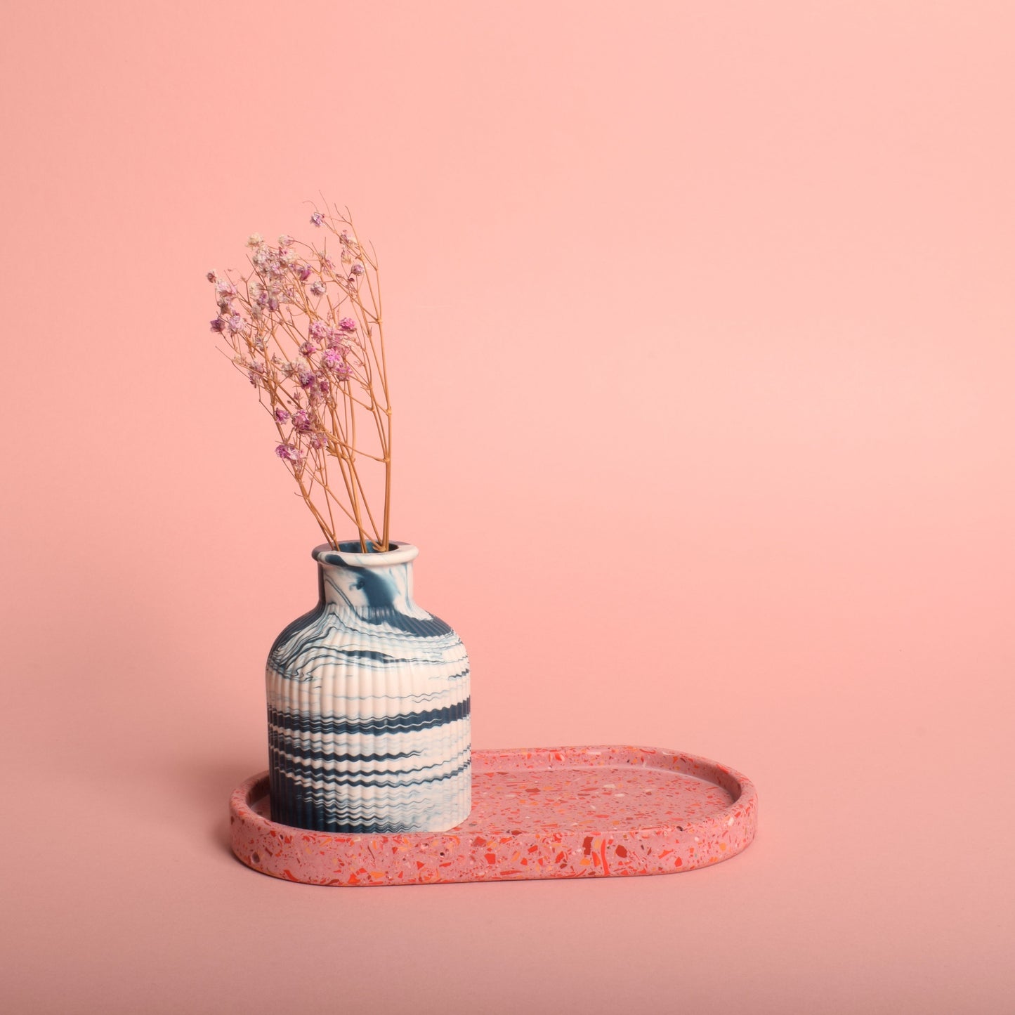 An oval pink terrazzo trinket tray holding a blue and white marble effect vase with dried flowers in it