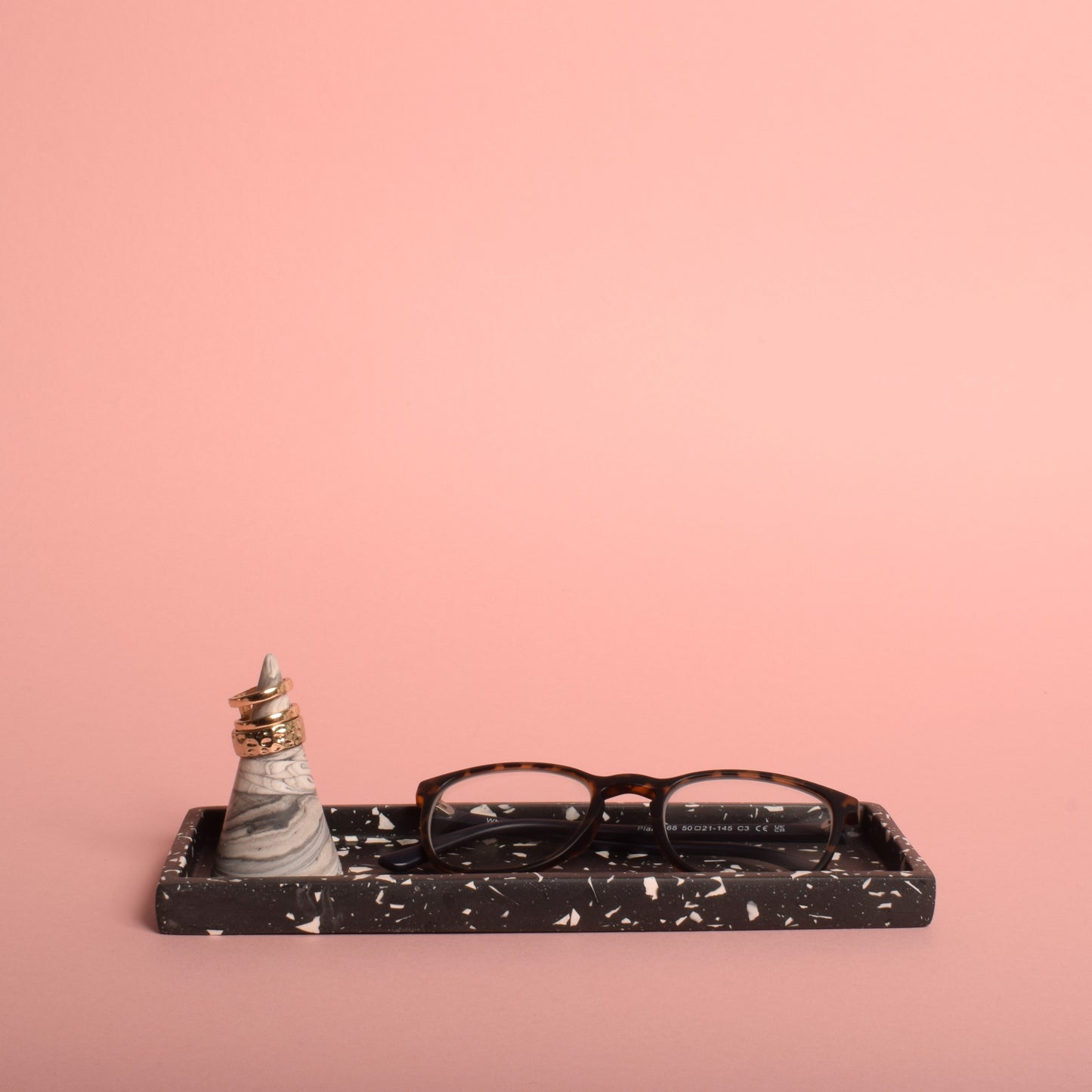 A rectangular black trinket tray with white terrazzo chips in it. The tray is holding a grey and white ring holder with rings on it, and a pair of glasses.