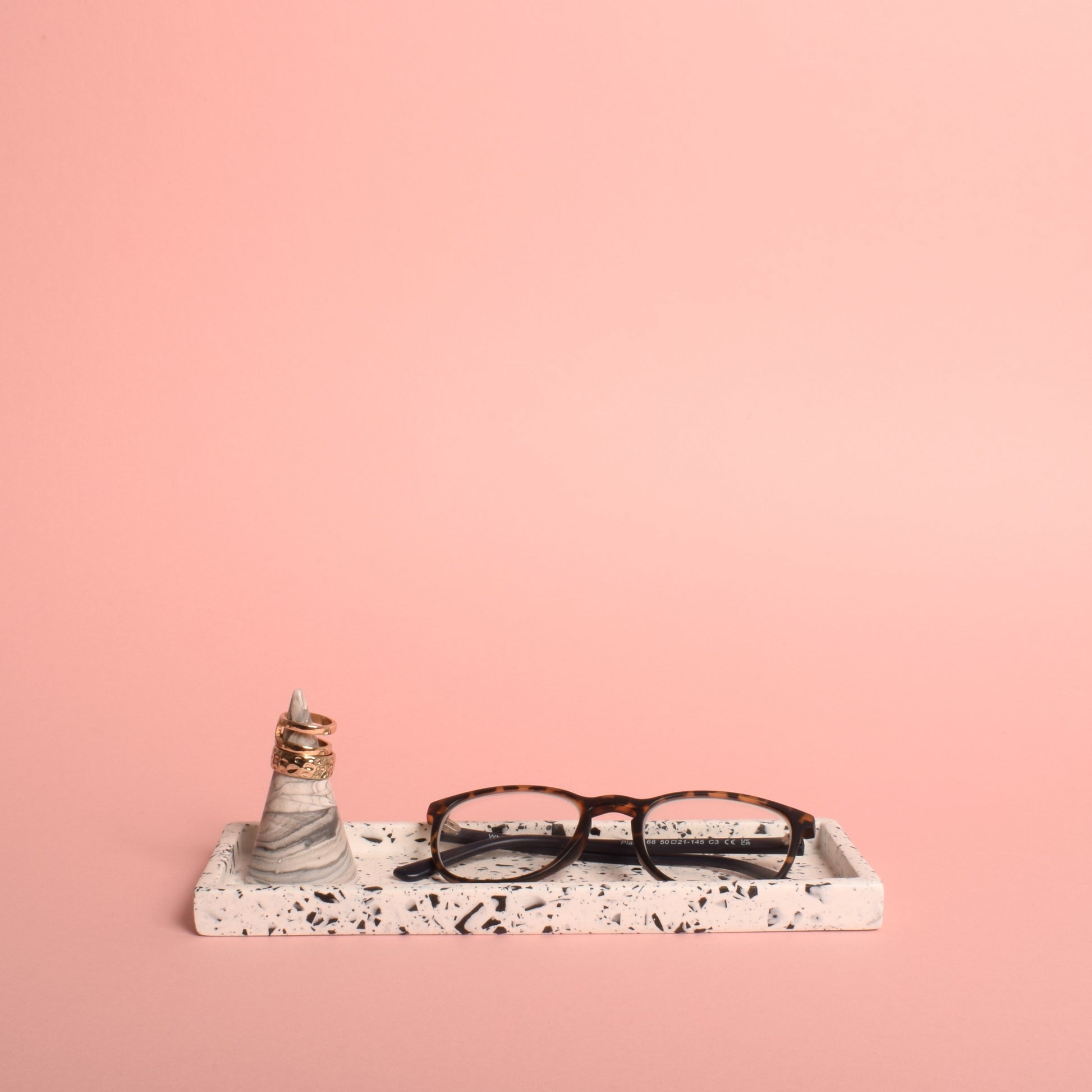 A rectangular white trinket tray with black terrazzo chips in it. The tray is holding a pair of glasses and a grey and white ring holder with gold rings on it.