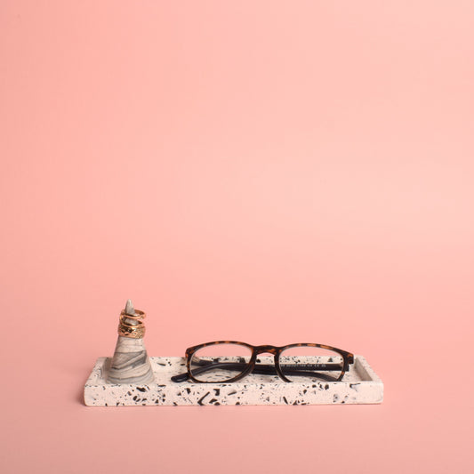 A rectangular white trinket tray with black terrazzo chips in it. The tray is holding a pair of glasses and a grey and white ring holder with gold rings on it.